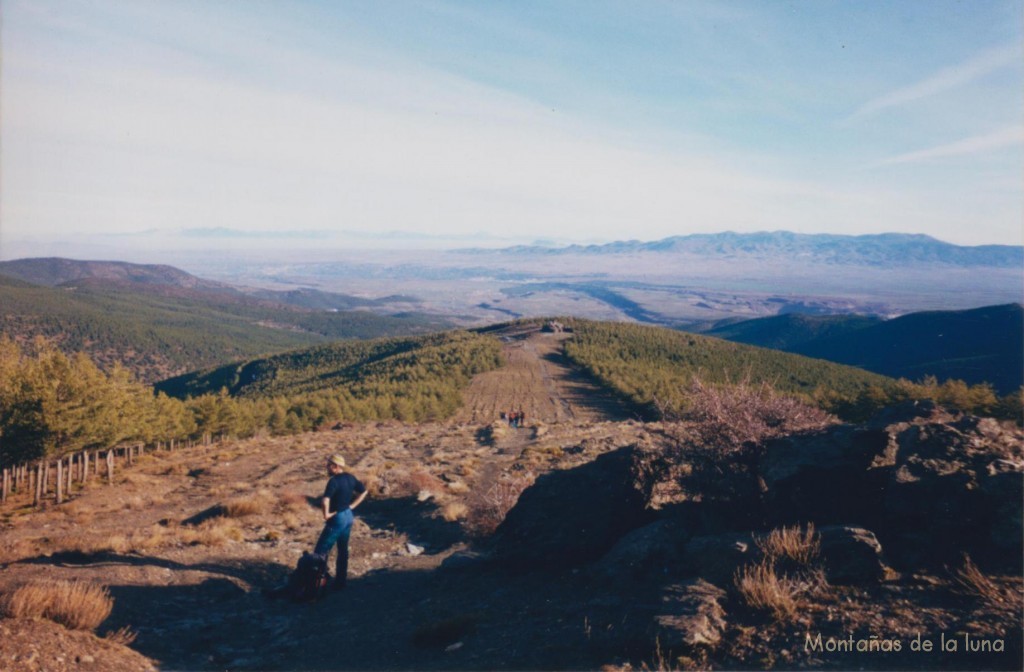 Delante Tomás en el cruce de caminos para seguir por la Loma de Enmedio, atrás queda el Refugio Postero Alto y abajo el llano de la Hoya de Guadix y Marquesado de Zenete con la Sierra de Baza a la derecha. Al fondo casi se percibe La Sagra en el centro, y el conjunto de las Sierras de Cazorla, Castril... a la izquierda